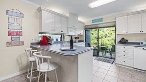 Une cuisine moderne avec des meubles blancs, un îlot central doté de tabourets de bar, une machine à café rouge et une vue sur la verdure à l’extérieur.