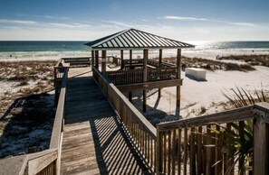 A wooden beach boardwalk leading to a covered gazebo with a metal roof, situated on a sandy beach with dunes in the background.