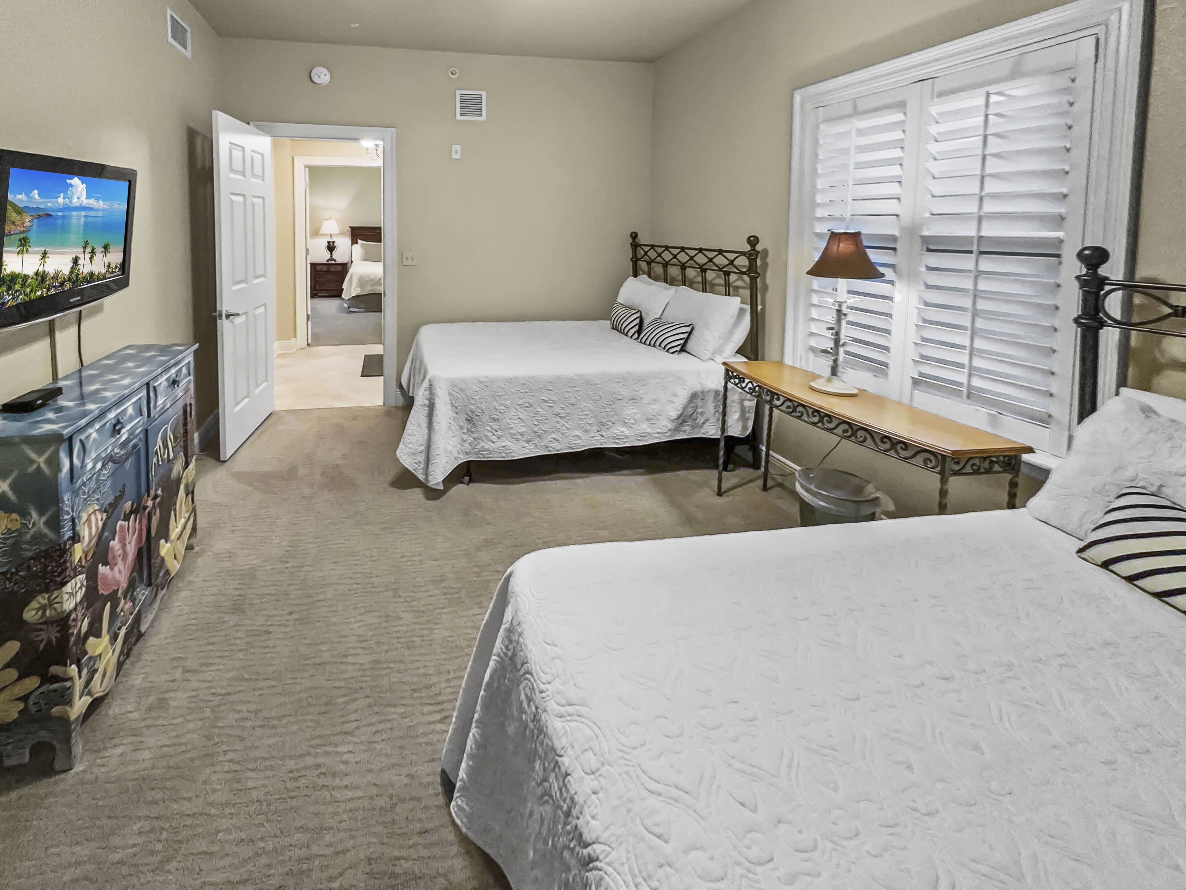 A bedroom with two beds, a desk, a TV, and a door leading to another room.