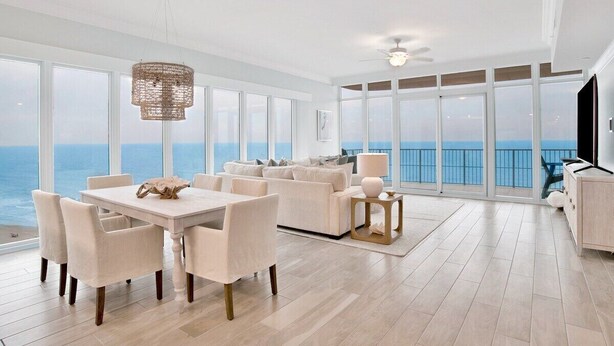 A spacious living area with a large window overlooking the ocean, a dining table with chairs, a sofa, and a ceiling fan. - Phoenix Gulf Tower 1704 | Spacious, luxurious, Newest OBA development! (Orange Beach)