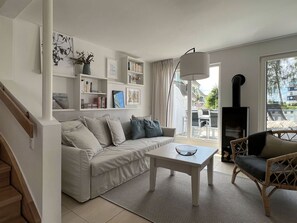 Ein modernes Wohnzimmer mit einer Couch, einem Couchtisch und einem Korbstuhl. Ein Bücherregal mit Büchern und ein Fernseher auf einem Schrank sind zu sehen. Eine Treppe ist links im Bild erkennbar.