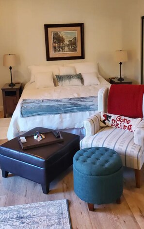 Un dormitorio con cama, mesita de noche, una silla, un pequeño otoman y una alfombra.
