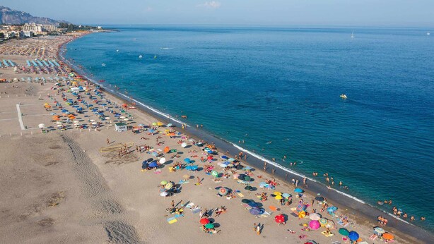Ein Strand mit vielen Sonnenschirmen und Liegestühlen, blauem Meer und ein paar Booten in der Ferne.