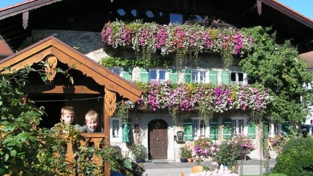 Une maison traditionnelle en bois, typique des Alpes, dotée d’un balcon orné de fleurs, d’une porte d’entrée et d’un petit jardin.