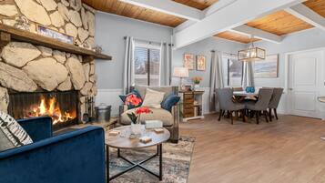 A living room with a stone fireplace, blue sofa, dining area with chairs, and a wooden ceiling.