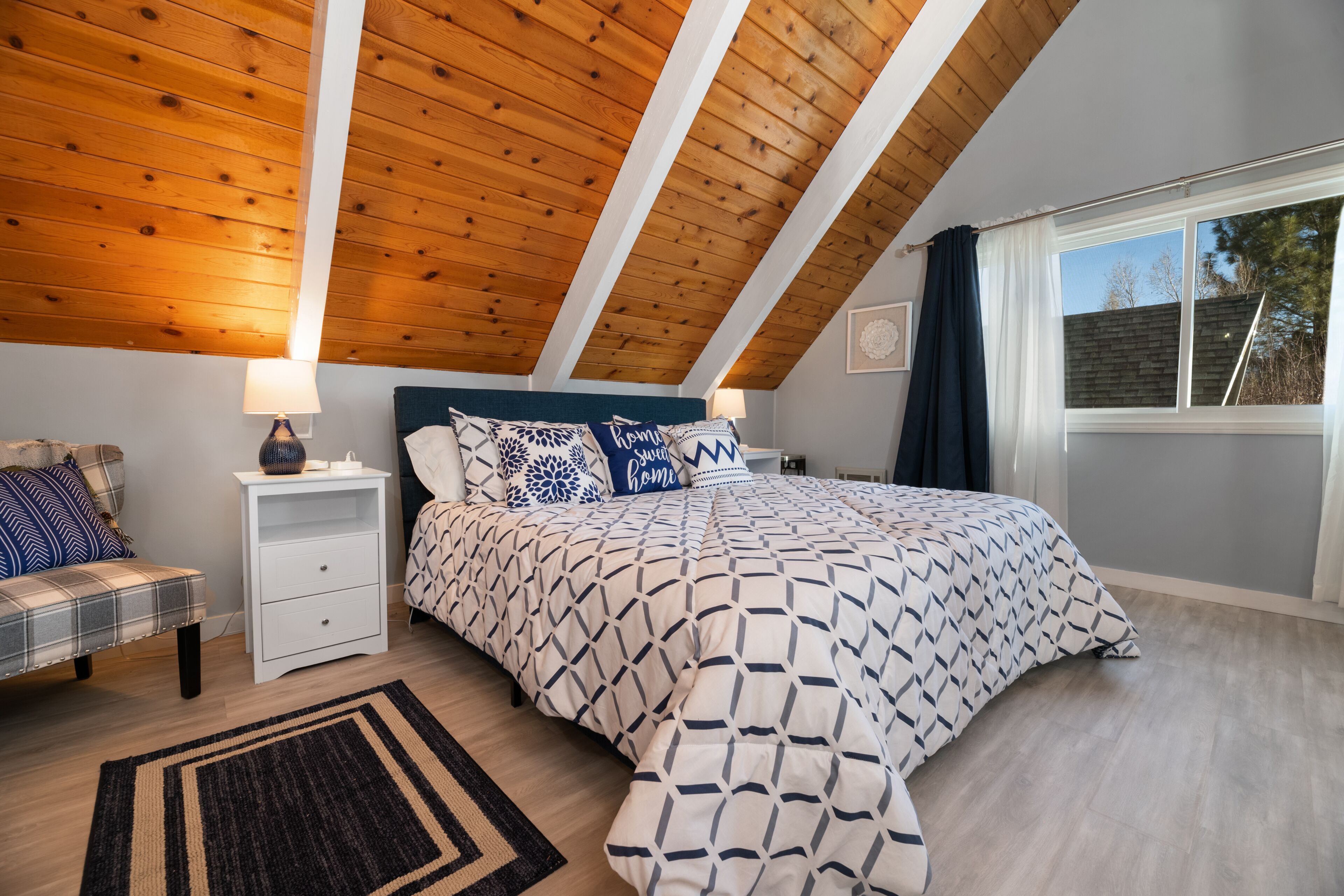 A bedroom with a sloped ceiling, a bed with patterned bedding, a nightstand, a bench, and a window with curtains.