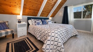 A bedroom with a sloped ceiling, a bed with patterned bedding, a nightstand, a bench, and a window with curtains. - Aqua Dreams (Big Bear City)