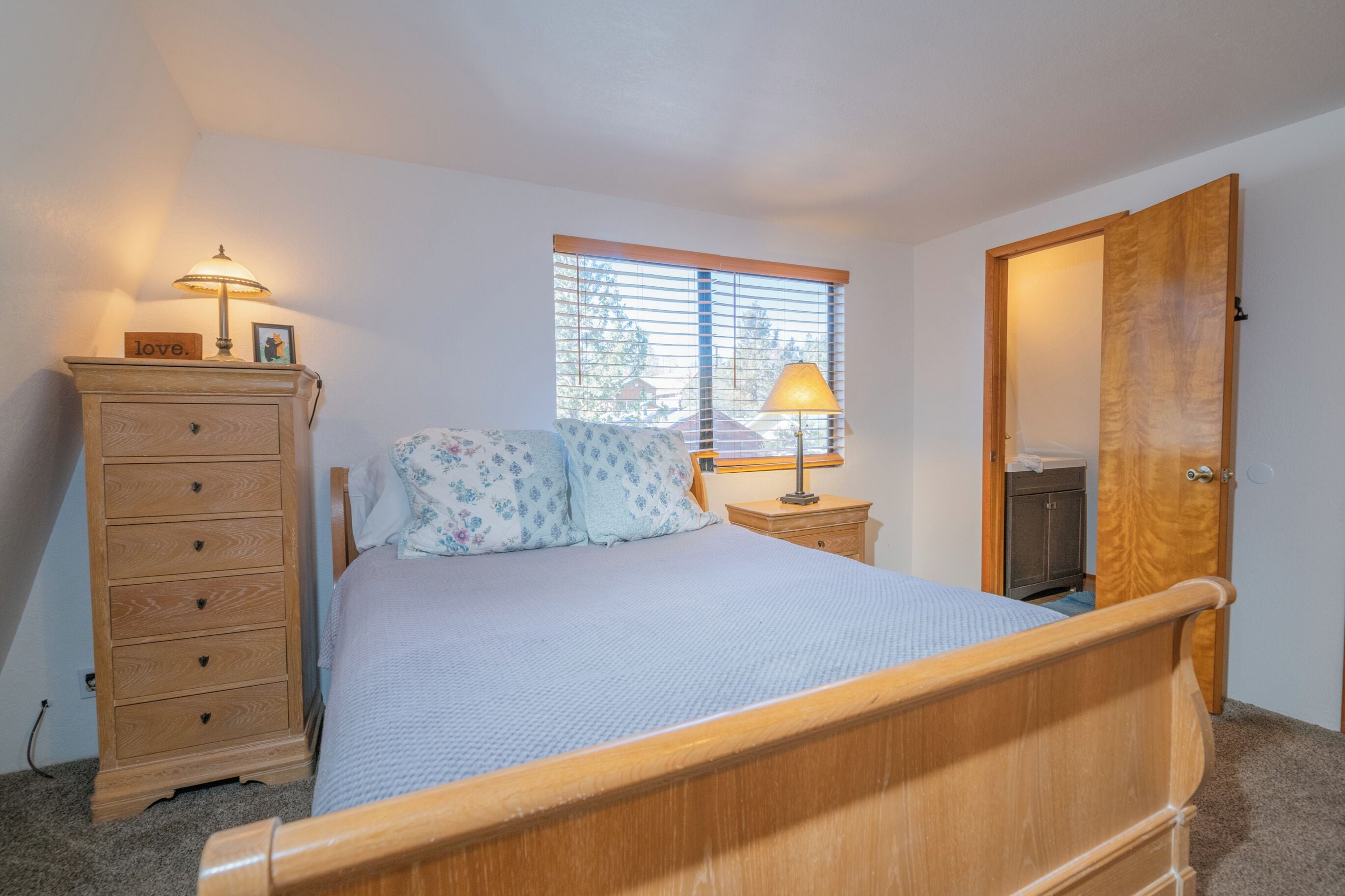 Un dormitorio con una cama de madera, un aparador, una mesita de noche con lámpara, una ventana con persianas y una puerta.
