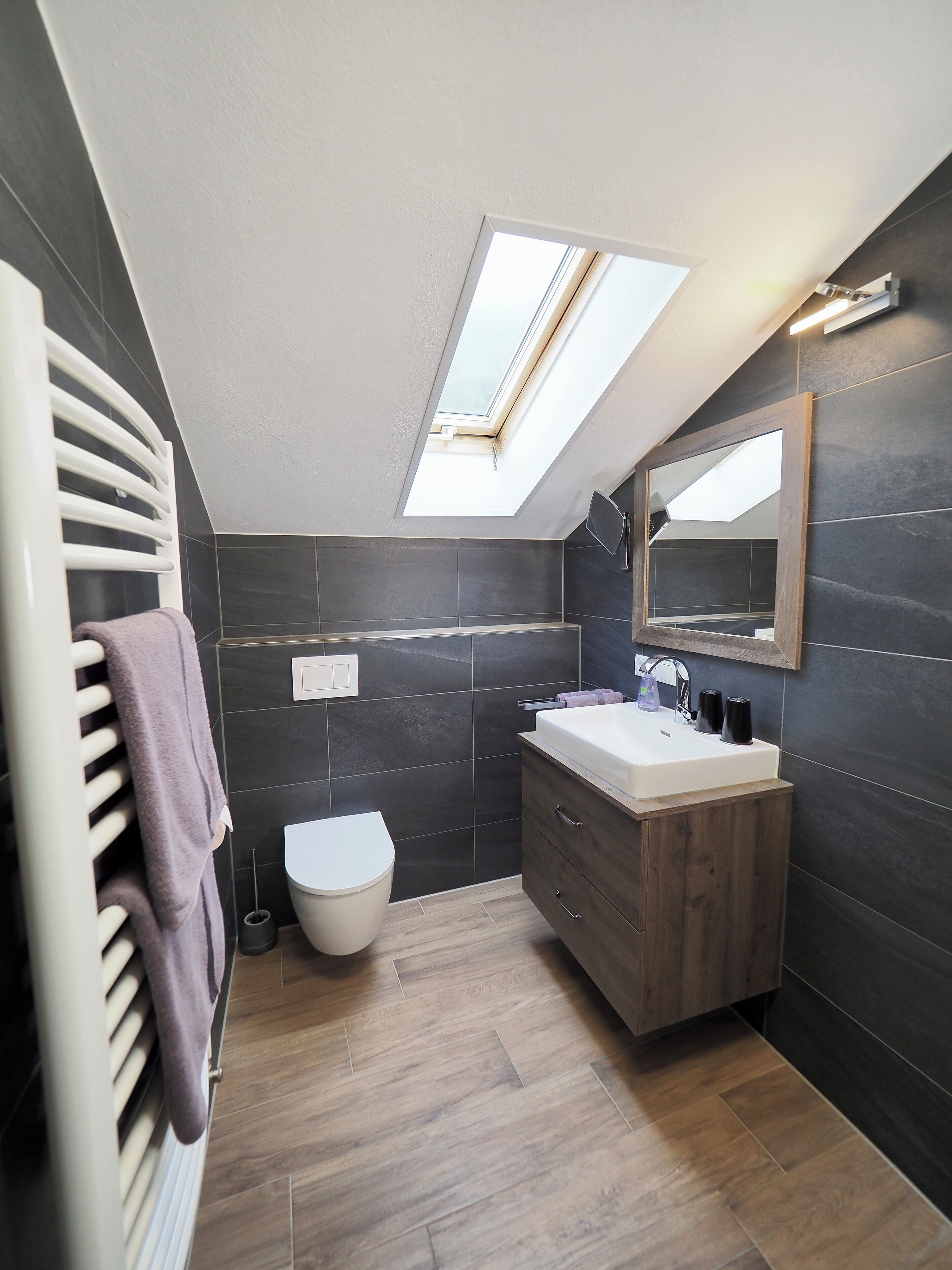 A modern bathroom with a skylight, dark tiles, a white toilet, a wooden vanity, and a towel rack.
