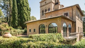 Un edificio storico con una torre, finestre ad arco e un giardino con siepi e un albero grande.