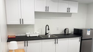 A compact kitchen with white cabinets, a black countertop, a stainless steel refrigerator, and a small wooden stool with a lamp. - Haven in the Prairies (2BR Suite) (Winnipeg)