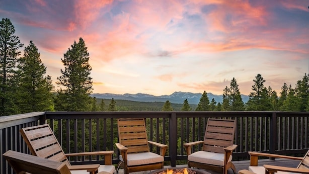 A deck with four wooden chairs and a central fire pit, surrounded by a wooden railing with a view of mountains and trees.