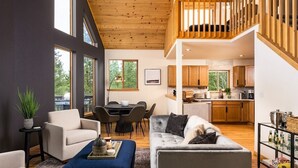 A modern living room with a high ceiling, wooden beams, a dining area, and a kitchen with wooden cabinets.
