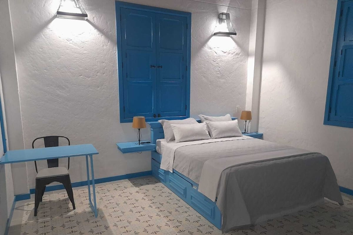 Un dormitorio con una cama, dos mesitas de noche, una silla y una ventana con contraventanas azules.