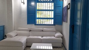 Una habitación con un sofá seccional blanco, una mesita pequeña blanca, un armario azul y una ventana con contraventanas azules.