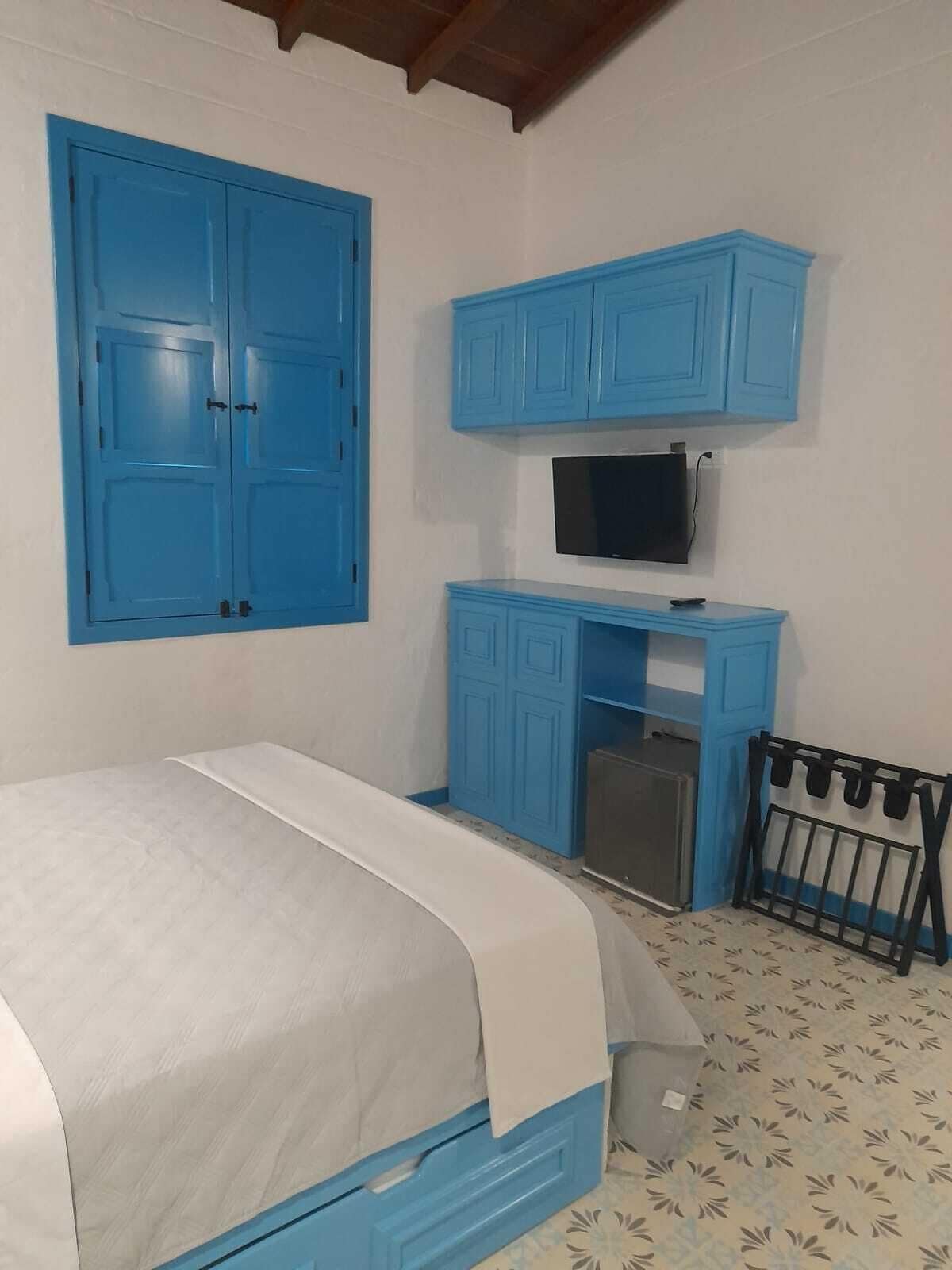 Quarto com cama, TV fixada em um móvel azul e um frigorífico pequeno.