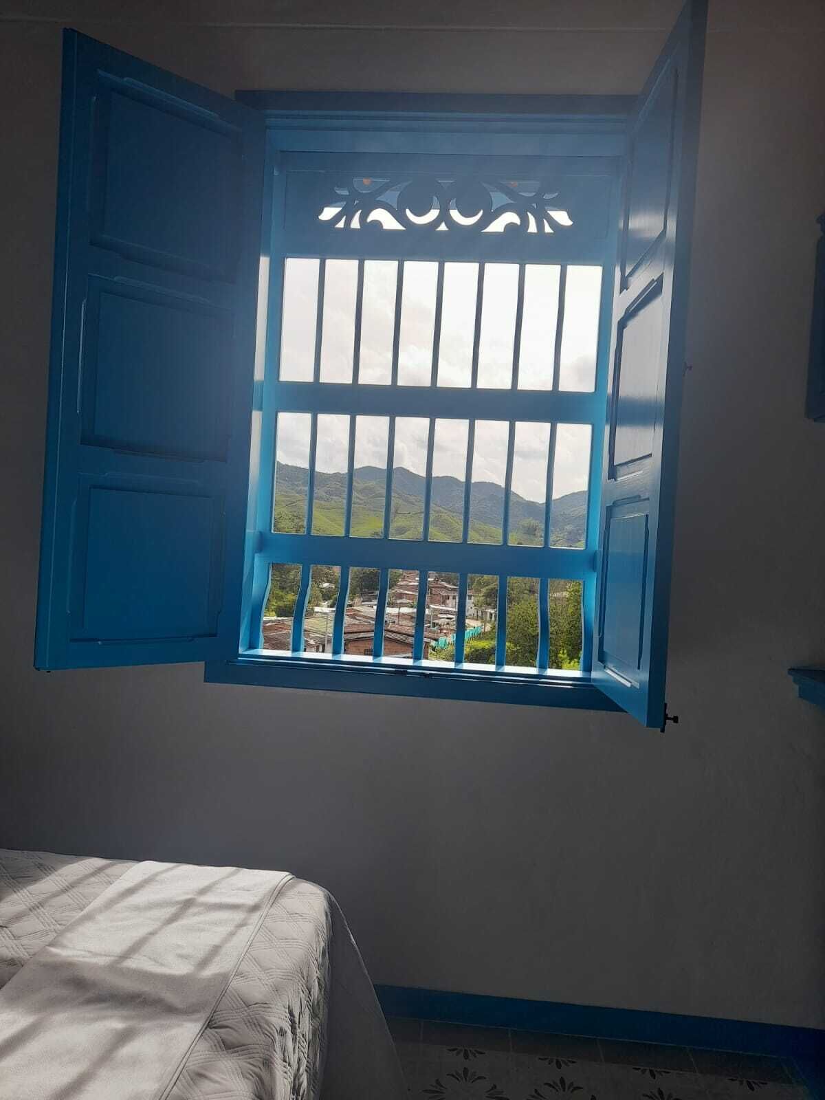Una habitación con cama, ventana con contraventanas azules y vista a un paisaje montañoso.