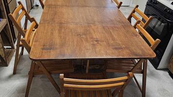 Een rechthoekige houten eettafel met zes stoelen, een keuken met een koelkast, fornuis en waterdispenser, en een raam met zonwering.