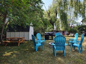 A backyard with a wooden picnic table, a grill, and blue Adirondack chairs.