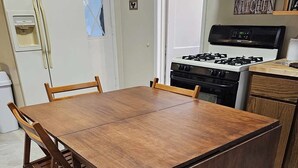 Een eetgedeelte met een houten tafel en stoelen, een keuken met fornuis en oven, en een koelkast.