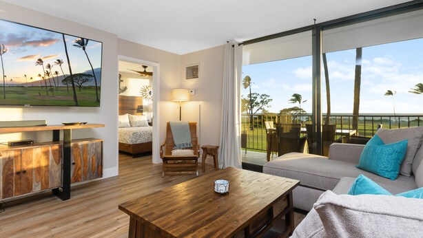 A modern living room with a wooden coffee table, a sofa with blue cushions, a TV mounted on the wall, and a view of the outdoors. - Modern & Elegant Oceanfront Vistas (Kihei)