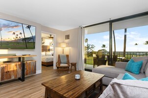 A modern living room with a wooden coffee table, a sofa with blue cushions, a TV mounted on the wall, and a view of the outdoors.