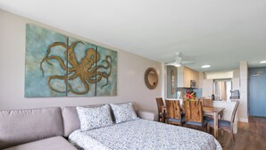 A modern bedroom with a large bed, a dining area with wooden furniture, and a kitchen in the background. - Modern & Elegant Oceanfront Vistas (Kihei)