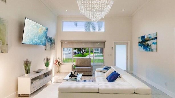 A modern living room with a white sofa, a flat-screen TV, a console table with decorative items, and a large chandelier. - Stylish Aventura Home With Pool on Intercostal (Ojus)