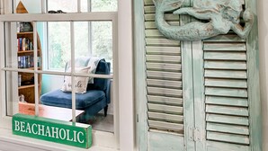 A room with a window featuring a 'BEACHAHOLIC' sign, a bookshelf, and a view of a blue chair and a pillow. - "High Tide Tranquility"- Beach w/ BBQ (Port Republic)