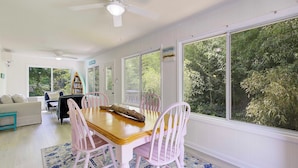 A dining area with a wooden table and pink chairs, large windows with a view of greenery, a ceiling fan, and a bookshelf. - "High Tide Tranquility"- Beach w/ BBQ (Port Republic)