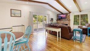 A spacious living room with a wooden floor, a brown leather sofa, a blue armchair, a dining table with chairs, and a TV mounted on a brick wall. - "High Tide Tranquility"- Beach w/ BBQ (Port Republic)