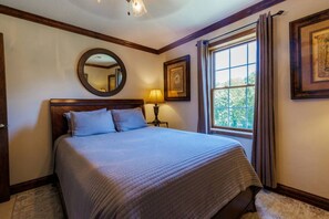 Una cama bien hecha con cabecera, una lámpara de noche, una ventana con cortinas y un cuadro en la pared.