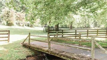 A rustic wooden bridge over a small stream in a park with a wooden fence and bench in the background.