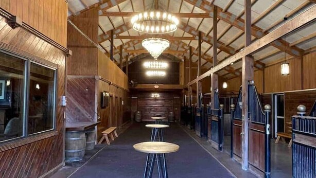 A spacious indoor area with a high ceiling, wooden beams, and a central chandelier. There are metal stanchions and a brick floor. - The Stables Cottage (Knoxville)