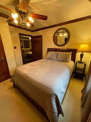 A bedroom with a large bed, a ceiling fan, a round mirror, and a nightstand with a lamp.
