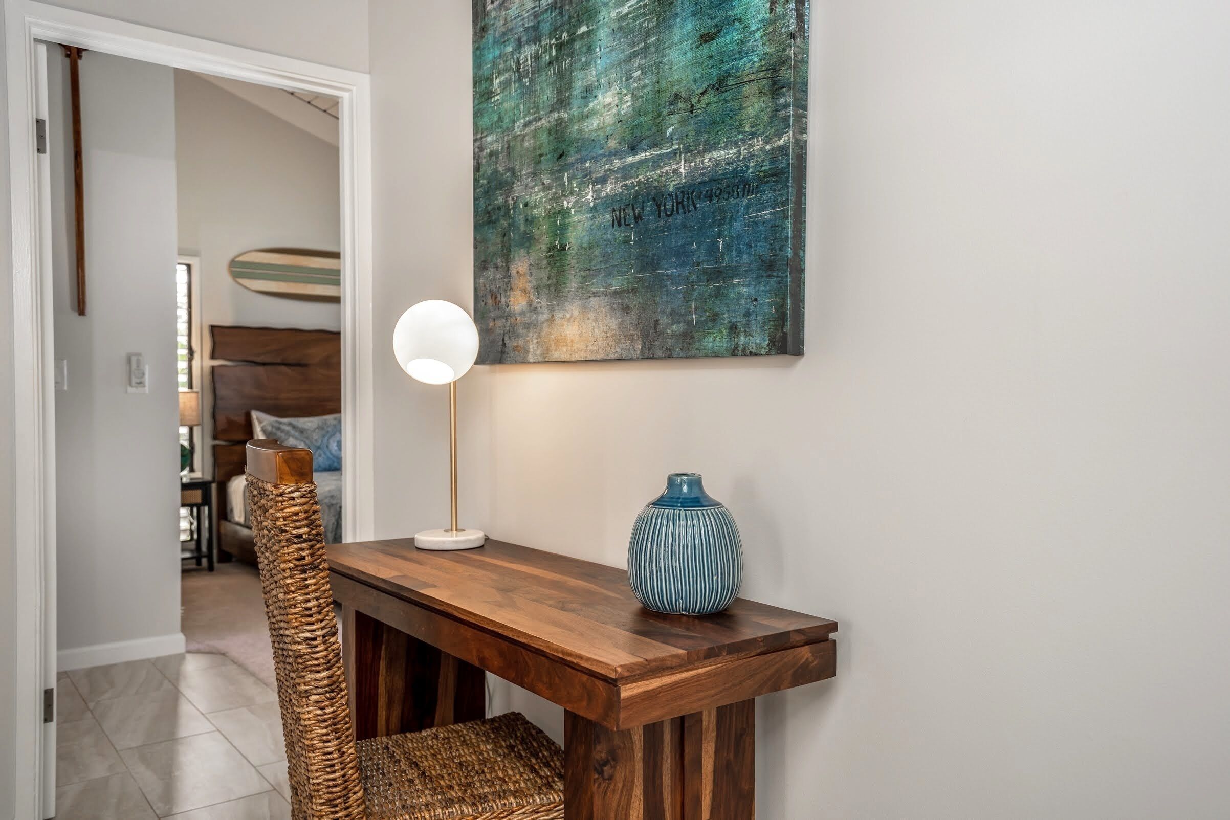 Une console en bois ornée d’un vase et d’une lampe, une chaise en osier et un mur sur lequel est accrochée une peinture à la texture particulière.