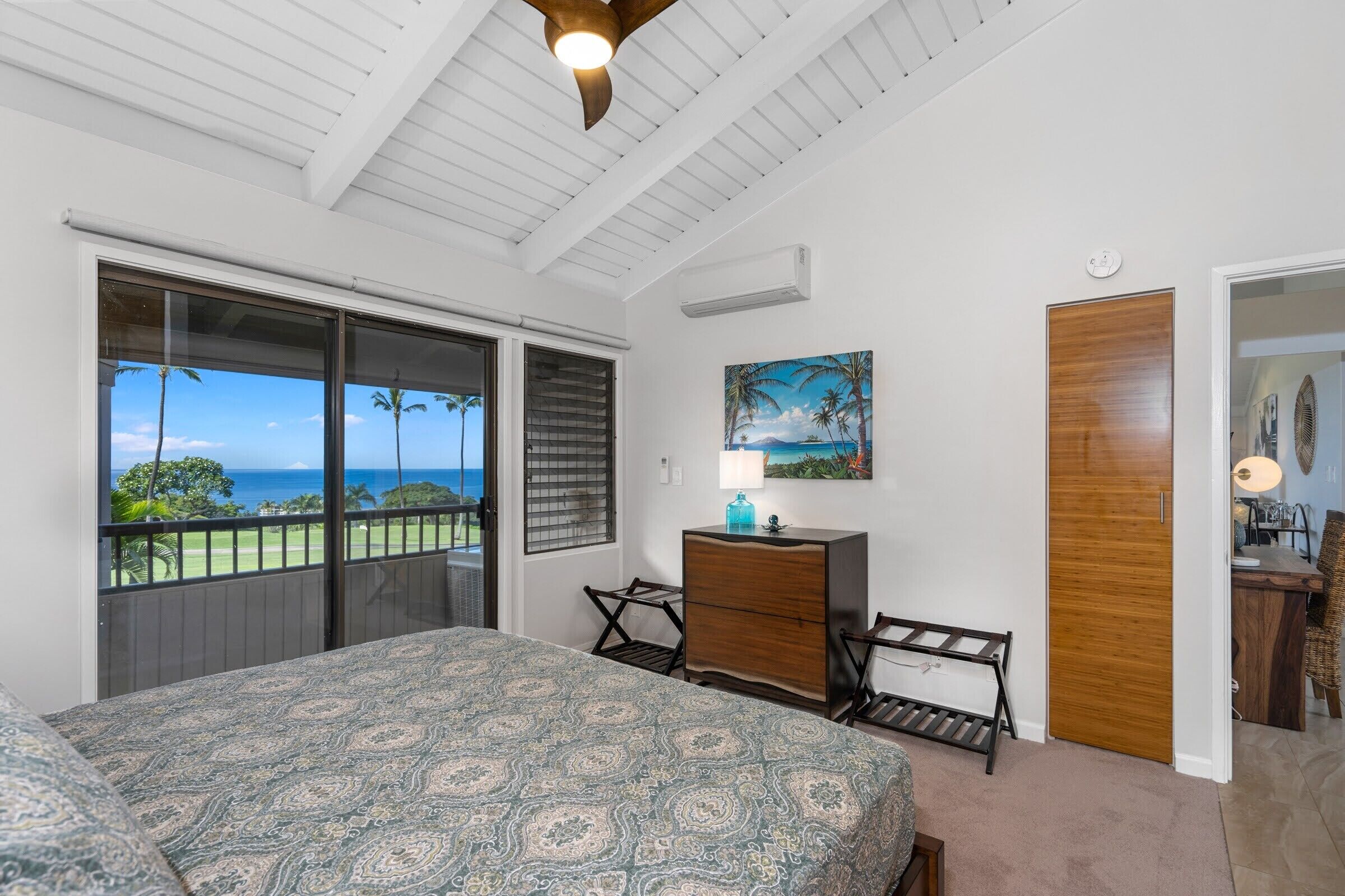 A bedroom with a large bed, a dresser, a desk, and a chair. There is a window with a view of the ocean and palm trees.