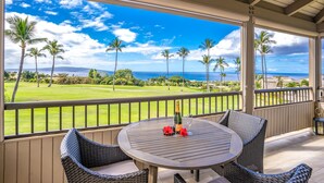 A covered outdoor area with a round table, wicker chairs, and a view of a golf course and ocean.