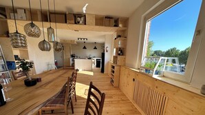 A modern kitchen with wooden cabinetry, a dining area with a long table and chairs, and a large window with a view of the outside. - Studio Loft Filmatelier (Hamburg)