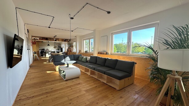 A modern living room with a sectional sofa, wooden flooring, large windows, and a kitchen in the background. - Studio Loft Filmatelier (Hamburg)