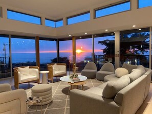 A modern living room with a large window overlooking a sunset, furnished with sofas, chairs, and a coffee table. - Ocean View Moss Beach Retreat (Moss Beach)