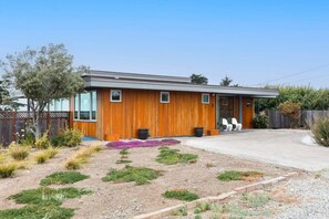 A modern single-story house with a wooden exterior, a front porch with chairs, and a landscaped garden with various plants and a gravel path. - Ocean View Moss Beach Retreat (Moss Beach)