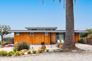 A modern two-story house with a wooden facade, a large palm tree, and a gravel driveway.