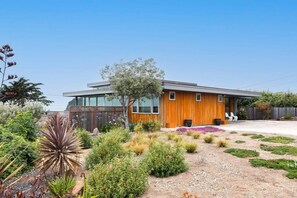 A modern house with a wooden exterior, a large glass window, and a well-maintained garden with various plants and shrubs. - Ocean View Moss Beach Retreat (Moss Beach)