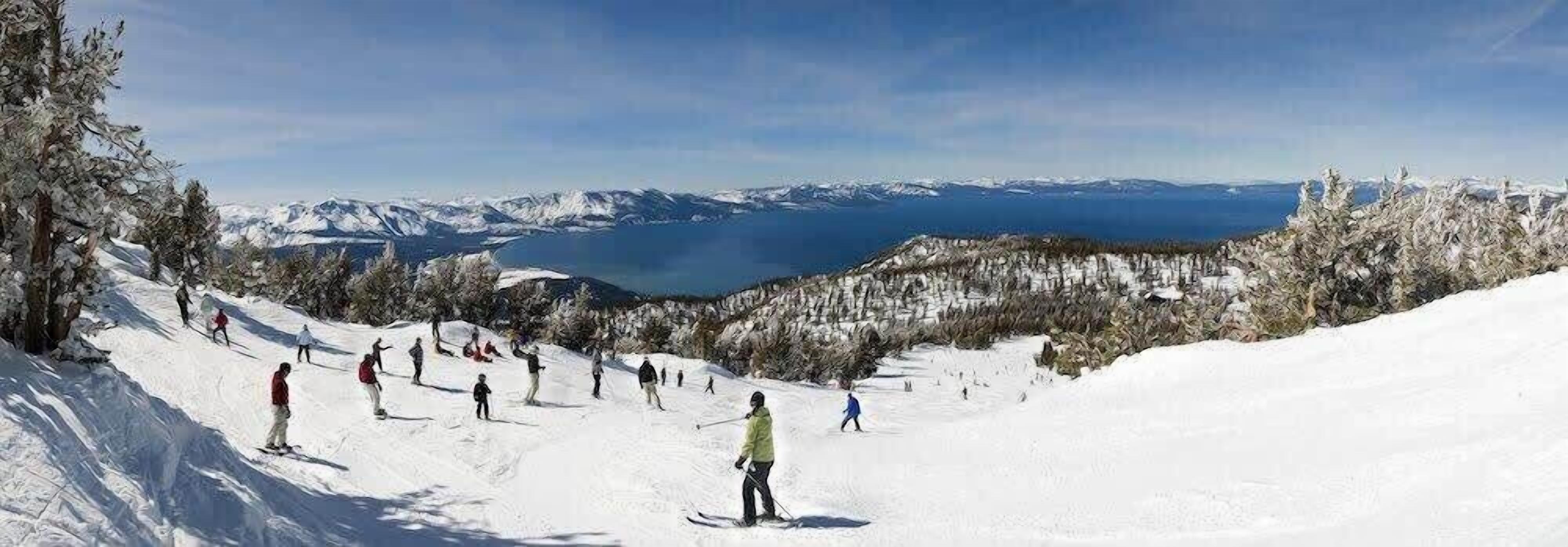 Pendiente nevada de una montaña con esquiadores y un lago al fondo.