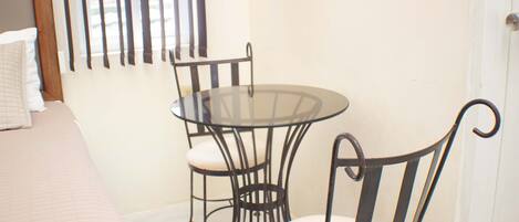 A room with a bed, a round glass table, two chairs, and a window with blinds.