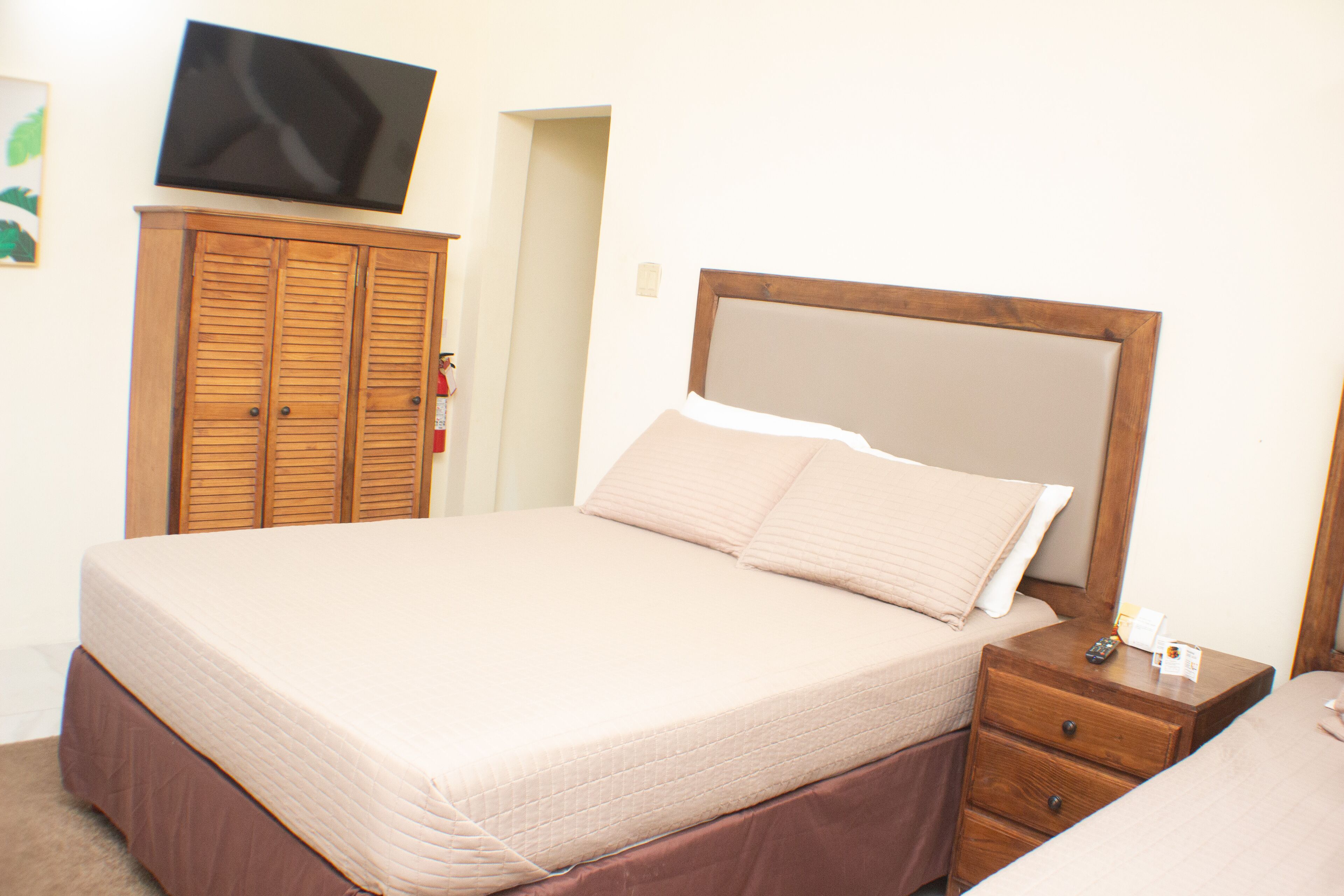 Una cama bien hecha con cabecera de madera, una mesita de noche con control remoto y un armario con puertas persianas.