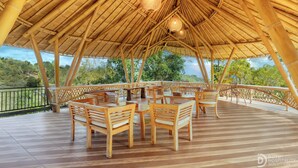 An outdoor bamboo-roofed area with wooden tables and chairs, a railing, and a view of greenery. - Tinggian Hill Retreat (Penida Island)