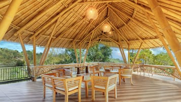 An outdoor bamboo-roofed area with wooden tables and chairs, a railing, and a view of greenery.