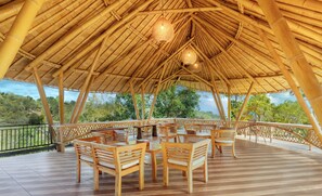 Un espacio al aire libre con techo de bambú, mesas y sillas de madera, barandilla y vista a follaje.